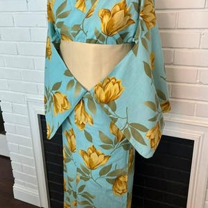 May include: A light blue kimono featuring a repeating pattern of large golden-yellow tulip-like flowers and green leaves. The garment has wide, flowing sleeves and is cinched at the waist with a solid, pale cream-colored obi sash belt.