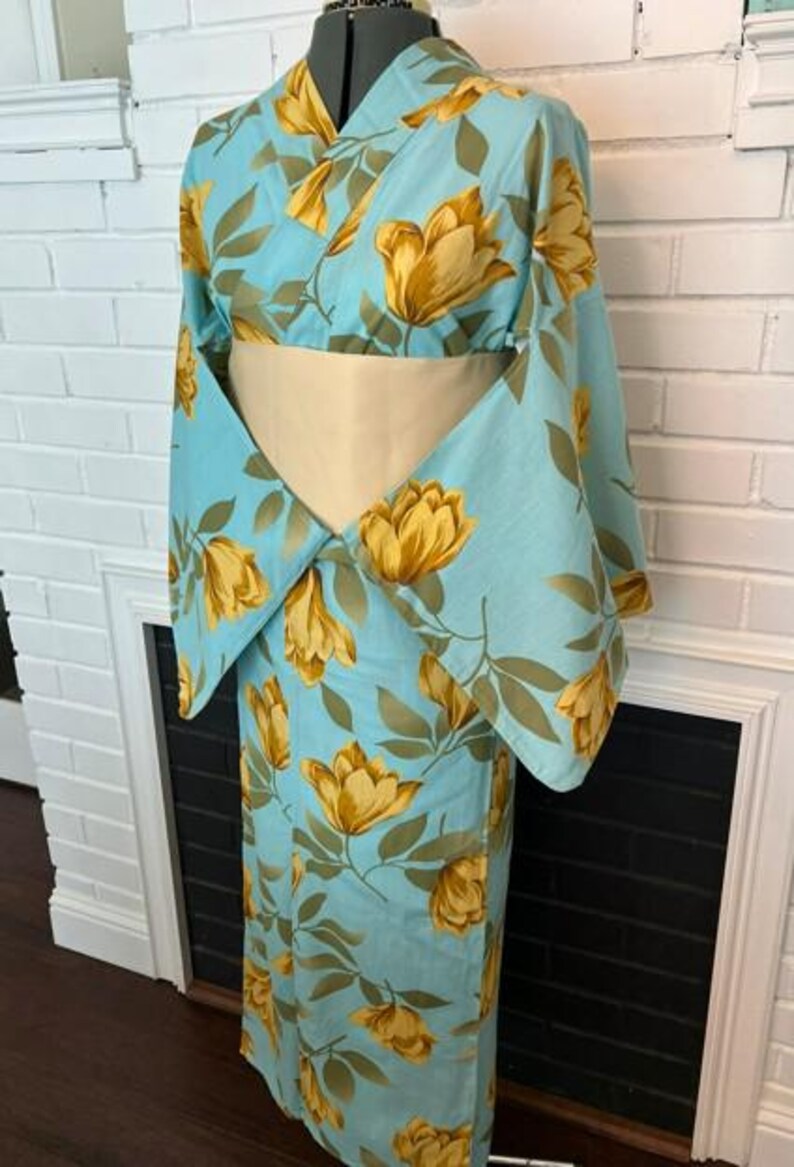 May include: A light blue kimono featuring a repeating pattern of large golden-yellow tulip-like flowers and green leaves. The garment has wide, flowing sleeves and is cinched at the waist with a solid, pale cream-colored obi sash belt.