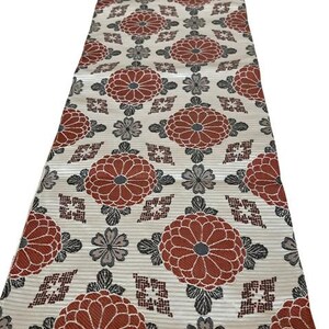 May include: A rectangular textile runner featuring a repeating pattern of large, rust-coloured chrysanthemum flowers interspersed with smaller grey floral motifs and geometric diamond shapes on a light cream-coloured background with a ribbed texture.