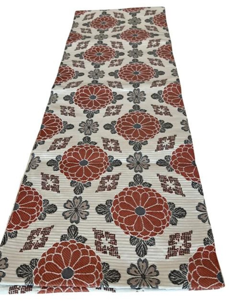 May include: A rectangular textile runner featuring a repeating pattern of large, rust-coloured chrysanthemum flowers interspersed with smaller grey floral motifs and geometric diamond shapes on a light cream-coloured background with a ribbed texture.