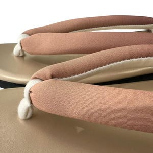 May include: Close-up of a pair of beige geta sandals. The sandals feature a light brown fabric strap with a white interior, attached to a beige platform. The sole is black. The image is set against a white background.