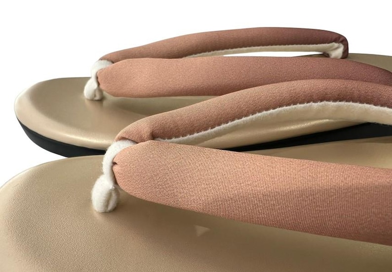 May include: Close-up of a pair of beige geta sandals. The sandals feature a light brown fabric strap with a white interior, attached to a beige platform. The sole is black. The image is set against a white background.