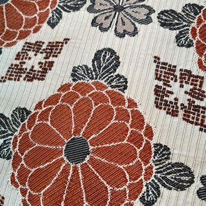 May include: A woven fabric featuring a repeating pattern of large, rust-orange chrysanthemum-like flowers with dark grey centres and leaves, interspersed with smaller grey floral motifs and geometric grid shapes on a light cream-coloured background.