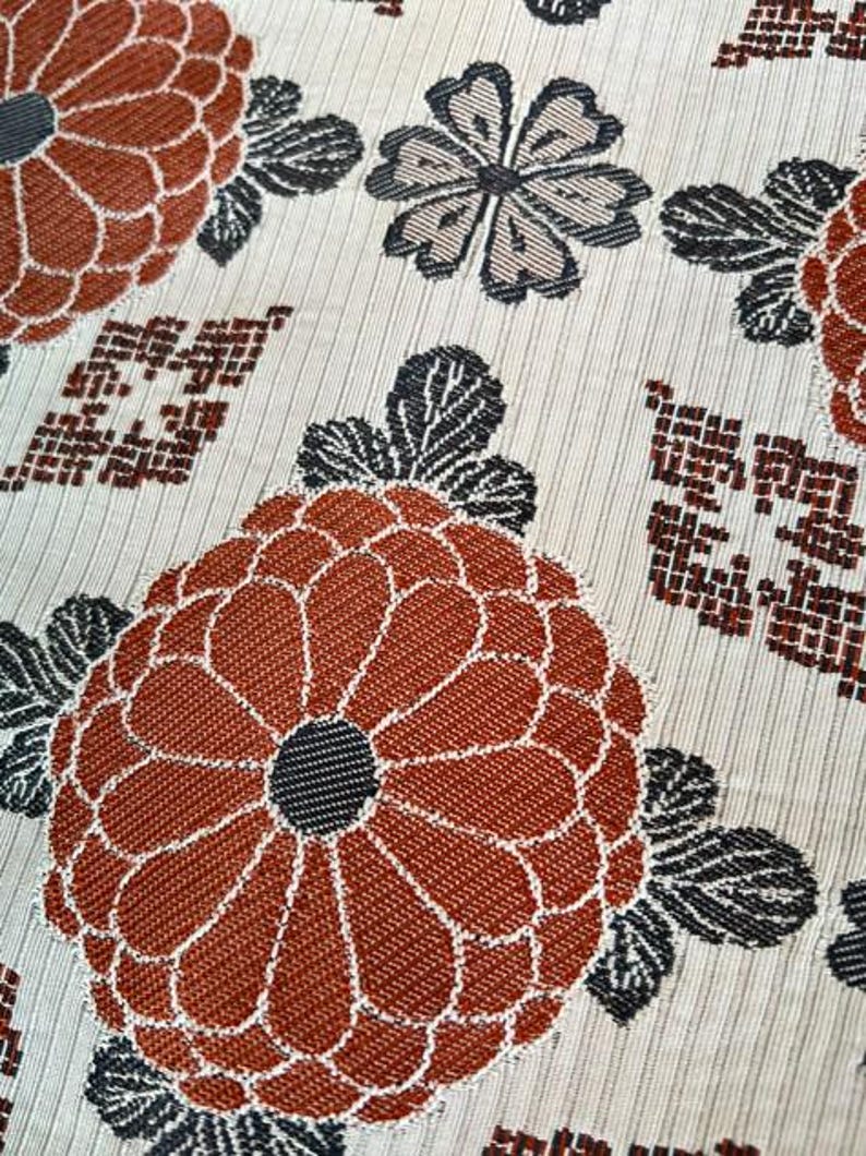 May include: A woven fabric featuring a repeating pattern of large, rust-orange chrysanthemum-like flowers with dark grey centres and leaves, interspersed with smaller grey floral motifs and geometric grid shapes on a light cream-coloured background.