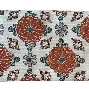 May include: A rectangular fabric piece featuring a repeating pattern of large rust-orange chrysanthemum-like flowers, smaller grey four-petaled blossoms, and dark brown geometric diamond shapes set against a light grey textured background.
