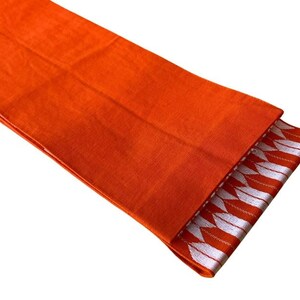 May include: A vibrant orange fabric runner featuring a textured weave. One end of the runner is folded over to reveal a decorative border pattern consisting of repeating white arrow-like shapes arranged in a horizontal row against an orange background.
