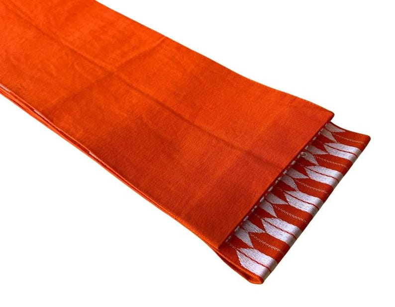 May include: A vibrant orange fabric runner featuring a textured weave. One end of the runner is folded over to reveal a decorative border pattern consisting of repeating white arrow-like shapes arranged in a horizontal row against an orange background.