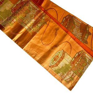 Op de afbeelding: Een oranje textiel met ingewikkelde patronen en ontwerpen. De stof heeft herhaalde geometrische vormen en decoratieve elementen in tinten groen, rood en goud. Het textiel heeft een glanzende, zijdeachtige uitstraling.