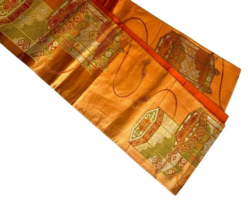 Op de afbeelding: Een oranje textiel met ingewikkelde patronen en ontwerpen. De stof heeft herhaalde geometrische vormen en decoratieve elementen in tinten groen, rood en goud. Het textiel heeft een glanzende, zijdeachtige uitstraling.