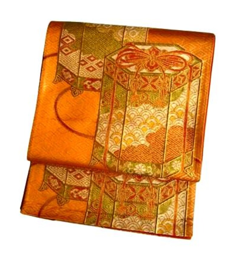 Op de afbeelding: Gevouwen oranje stof met een gedetailleerd patroon. Het ontwerp omvat geometrische vormen, een grote zeshoekige doos met een strik en golfachtige patronen. Gouden en groene accenten versterken de algehele esthetiek. De stof lijkt zijde of een vergelijkbaar materiaal te zijn.