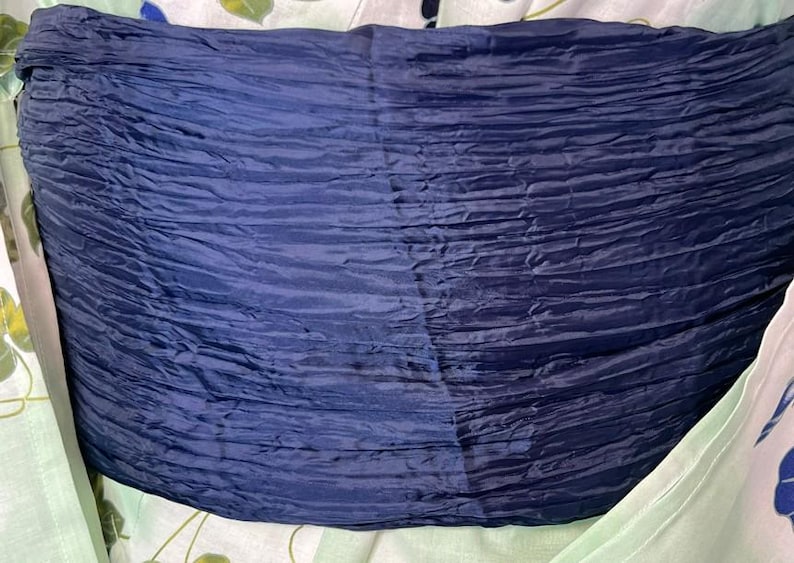 Op de afbeelding: Een brede, donker marineblauwe sjerp met een zwaar gekreukelde, horizontale textuur. De stof heeft een subtiele glans en is gewikkeld rond een lichtgekleurd kledingstuk dat een vaag, lichtgroen en blauw bloemenpatroon op het oppervlak toont.