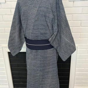 Może przedstawiać: Kimono w ciemnoniebiesko-biały wz&oacute;r z drobną geometryczną fakturą na całej tkaninie. Ubi&oacute;r posiada szerokie rękawy oraz jednolity, ciemnogranatowy pas obi owinięty wok&oacute;ł talii, ozdobiony pojedynczą, cienką, poziomą białą linią biegnącą przez środek.