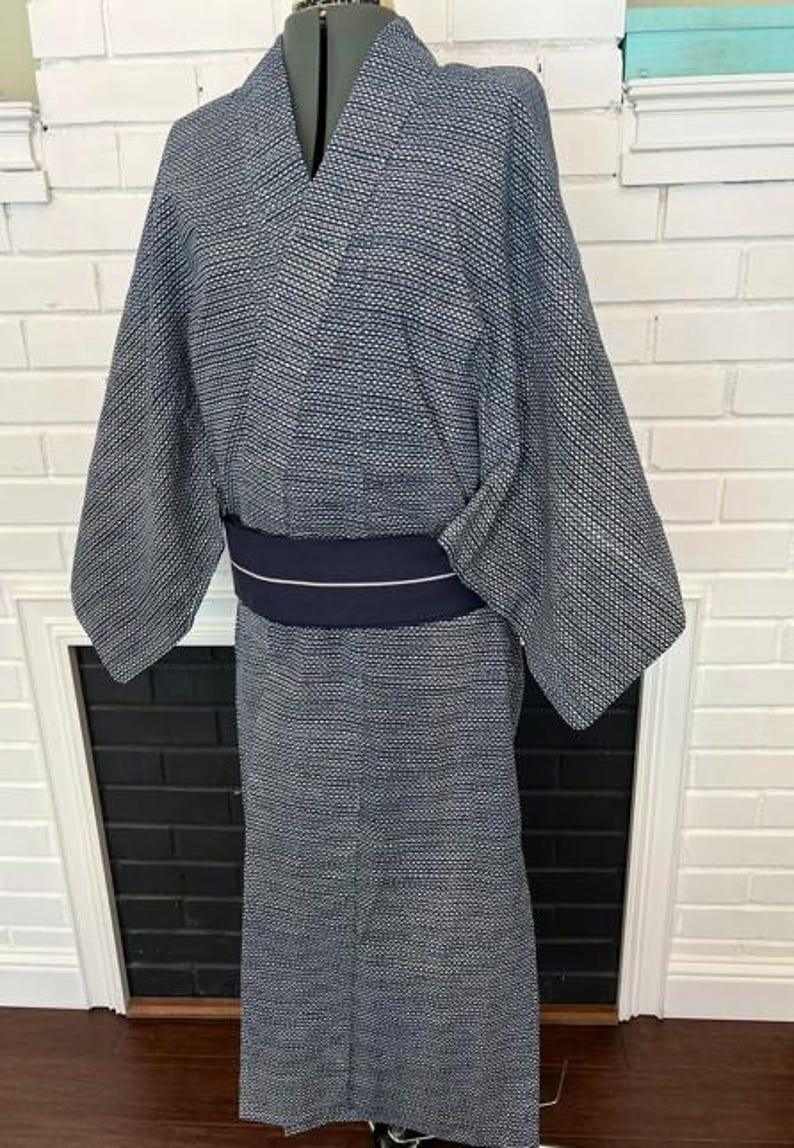 Może przedstawiać: Kimono w ciemnoniebiesko-biały wz&oacute;r z drobną geometryczną fakturą na całej tkaninie. Ubi&oacute;r posiada szerokie rękawy oraz jednolity, ciemnogranatowy pas obi owinięty wok&oacute;ł talii, ozdobiony pojedynczą, cienką, poziomą białą linią biegnącą przez środek.