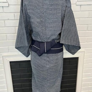 Może przedstawiać: Kimono w ciemnoniebiesko-biały wz&oacute;r z teksturowanym geometrycznym splotem na całej tkaninie. Ubi&oacute;r jest ściągnięty w talii jednolitym, ciemnogranatowym pasem obi, kt&oacute;ry zawiera cienki, biały detal w postaci poziomego paska.
