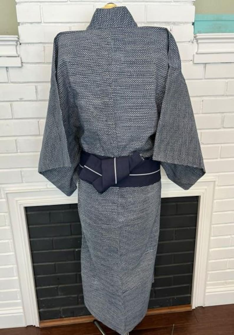 Może przedstawiać: Kimono w ciemnoniebiesko-biały wz&oacute;r z teksturowanym geometrycznym splotem na całej tkaninie. Ubi&oacute;r jest ściągnięty w talii jednolitym, ciemnogranatowym pasem obi, kt&oacute;ry zawiera cienki, biały detal w postaci poziomego paska.