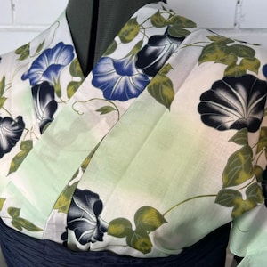 Op de afbeelding: Een yukata met een witte basis en een lichtgroen kleurverloop, versierd met een bloemenpatroon van blauwe en zwarte ochtendgloriebloemen en groene bladeren. Het kledingstuk heeft een overslagmodel met een brede kraag en een donkerblauwe obi-ceintuur.