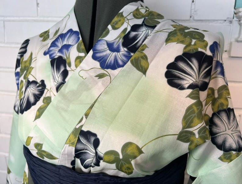 Op de afbeelding: Een yukata met een witte basis en een lichtgroen kleurverloop, versierd met een bloemenpatroon van blauwe en zwarte ochtendgloriebloemen en groene bladeren. Het kledingstuk heeft een overslagmodel met een brede kraag en een donkerblauwe obi-ceintuur.