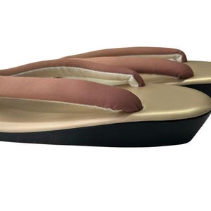 May include: A pair of traditional Japanese geta sandals. The sandals feature a gold-colored platform with a black base and brown fabric straps. The thong straps are padded for comfort. The geta are designed for outdoor wear.