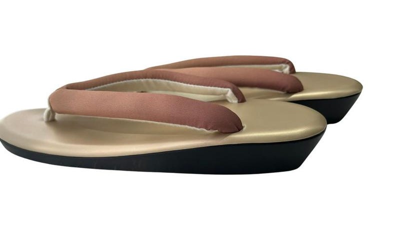 May include: A pair of traditional Japanese geta sandals. The sandals feature a gold-colored platform with a black base and brown fabric straps. The thong straps are padded for comfort. The geta are designed for outdoor wear.