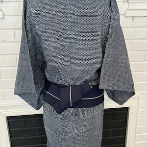 Może przedstawiać: Niebiesko-białe kimono we wzory z teksturowanym, geometrycznym splotem na całej tkaninie. Ubi&oacute;r jest ściągnięty w talii jednolitym, ciemnogranatowym pasem obi, kt&oacute;ry posiada pojedynczy, cienki detal w postaci pionowego białego paska.