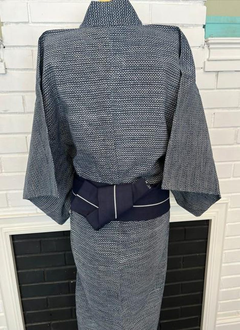 Może przedstawiać: Niebiesko-białe kimono we wzory z teksturowanym, geometrycznym splotem na całej tkaninie. Ubi&oacute;r jest ściągnięty w talii jednolitym, ciemnogranatowym pasem obi, kt&oacute;ry posiada pojedynczy, cienki detal w postaci pionowego białego paska.