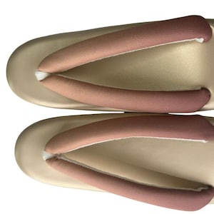 May include: A pair of traditional Japanese geta sandals. The sandals have a light beige base with two straps in a gradient of pink and brown. The thong straps are padded for comfort. The sandals are designed for outdoor wear.