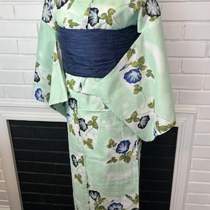 Op de afbeelding: Een licht mintgroene kimono met een herhalend patroon van blauwe en zwarte ochtendgloriebloemen met groene bladeren. Het kledingstuk is in de taille ingesnoerd met een brede, getextureerde donkerblauwe obi-ceintuur voor een gestructureerd silhouet.