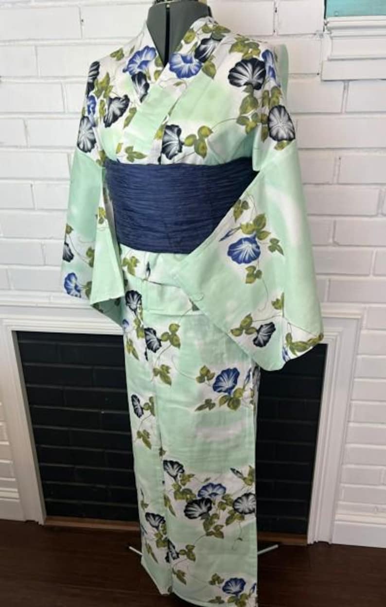 Op de afbeelding: Een licht mintgroene kimono met een herhalend patroon van blauwe en zwarte ochtendgloriebloemen met groene bladeren. Het kledingstuk is in de taille ingesnoerd met een brede, getextureerde donkerblauwe obi-ceintuur voor een gestructureerd silhouet.