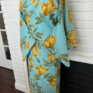 May include: A light blue kimono featuring a repeating pattern of large yellow flowers and green leaves. The garment has wide sleeves, a wrap-front closure, and a structured collar, displayed on a mannequin to show the full length and drape.