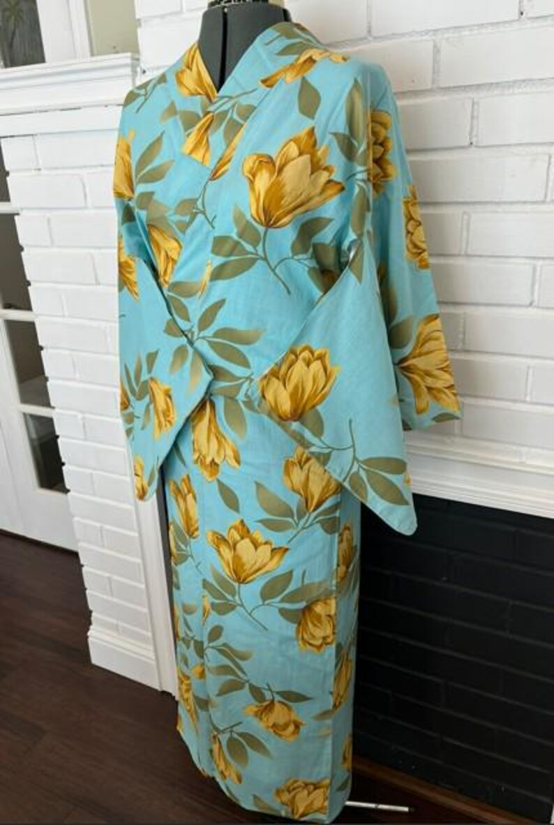 May include: A light blue kimono featuring a repeating pattern of large yellow flowers and green leaves. The garment has wide sleeves, a wrap-front closure, and a structured collar, displayed on a mannequin to show the full length and drape.