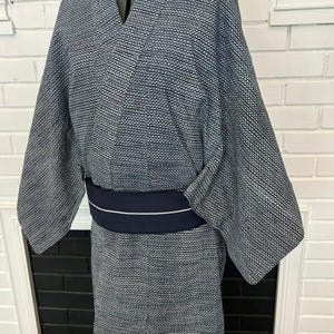 Może przedstawiać: Kimono w ciemnoniebiesko-biały wz&oacute;r z drobną geometryczną kratką na całej powierzchni tkaniny. Ubi&oacute;r jest przewiązany w talii jednolitym, ciemnogranatowym pasem obi, kt&oacute;ry posiada detal w postaci pojedynczego, cienkiego białego paska.