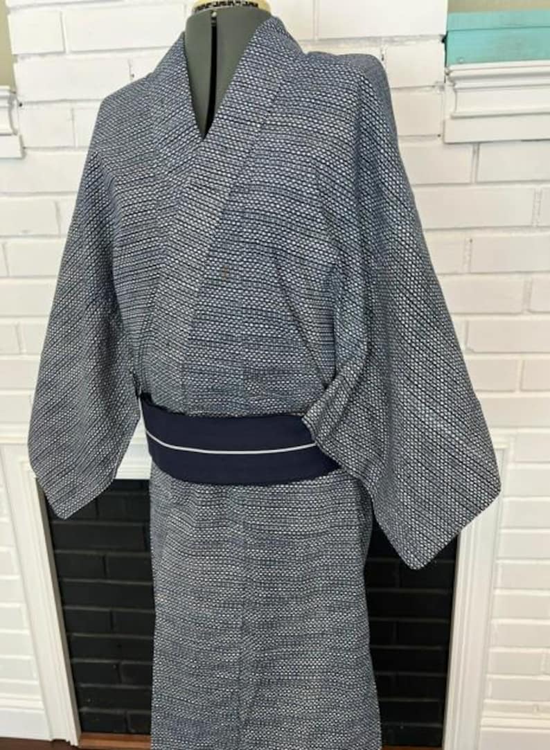 Może przedstawiać: Kimono w ciemnoniebiesko-biały wz&oacute;r z drobną geometryczną kratką na całej powierzchni tkaniny. Ubi&oacute;r jest przewiązany w talii jednolitym, ciemnogranatowym pasem obi, kt&oacute;ry posiada detal w postaci pojedynczego, cienkiego białego paska.