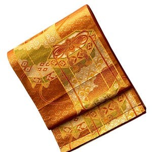 Op de afbeelding: Gevouwen oranje en gouden kimono met ingewikkelde geometrische en bloemmotieven. De stof heeft een herhalend ruitpatroon en een centraal paneel met een strikmotief. De randen zijn afgewerkt in een donkerdere oranje kleur.
