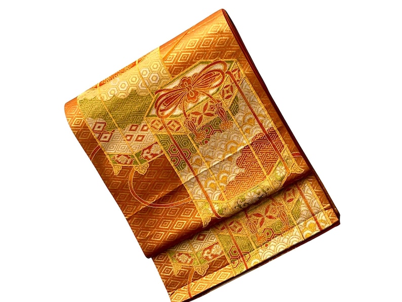Op de afbeelding: Gevouwen oranje en gouden kimono met ingewikkelde geometrische en bloemmotieven. De stof heeft een herhalend ruitpatroon en een centraal paneel met een strikmotief. De randen zijn afgewerkt in een donkerdere oranje kleur.