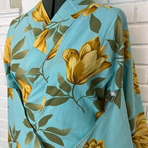 May include: A light blue kimono robe featuring a repeating pattern of large golden-yellow tulip-like flowers and muted olive green leaves. The garment has a wrap-front design with a wide collar and long sleeves, displayed on a mannequin torso.
