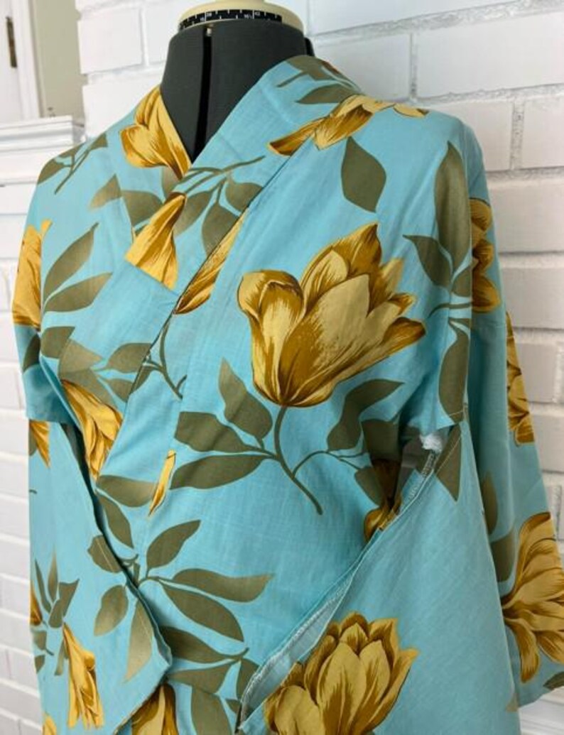 May include: A light blue kimono robe featuring a repeating pattern of large golden-yellow tulip-like flowers and muted olive green leaves. The garment has a wrap-front design with a wide collar and long sleeves, displayed on a mannequin torso.