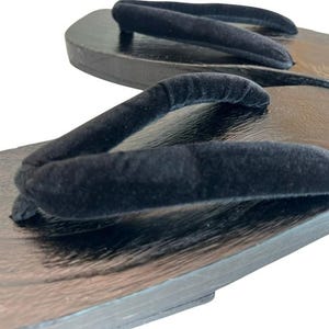 May include: A pair of geta sandals featuring dark brown wooden platforms with a polished finish and black velvet thong straps. The footwear has a flat, rectangular base design with a visible wood grain texture on the surface of the footbed.