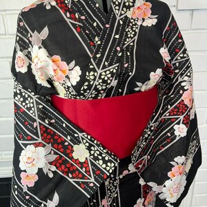 May include: A black kimono featuring a floral pattern with white and pink blossoms, accented by red leaf motifs and geometric white lines. The garment is cinched at the waist with a wide, solid red obi sash, showcasing wide sleeves and a traditional wrap-style front.