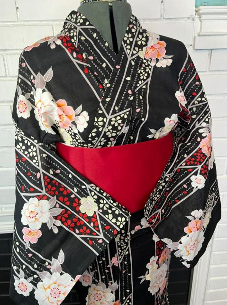 May include: A black kimono featuring a floral pattern with white and pink blossoms, accented by red leaf motifs and geometric white lines. The garment is cinched at the waist with a wide, solid red obi sash, showcasing wide sleeves and a traditional wrap-style front.