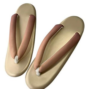 May include: A pair of traditional Japanese geta sandals. The sandals have a light beige base with brown and beige straps. The straps are padded for comfort. The geta sandals are designed to be worn with traditional Japanese clothing.