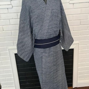 Może przedstawiać: Kimono w ciemnoniebiesko-biały wz&oacute;r z drobną geometryczną teksturą na całej tkaninie. Ubi&oacute;r posiada szerokie rękawy, dekolt w serek oraz jednolity ciemnogranatowy pas obi zawiązany w talii z cienkim białym poziomym paskiem.
