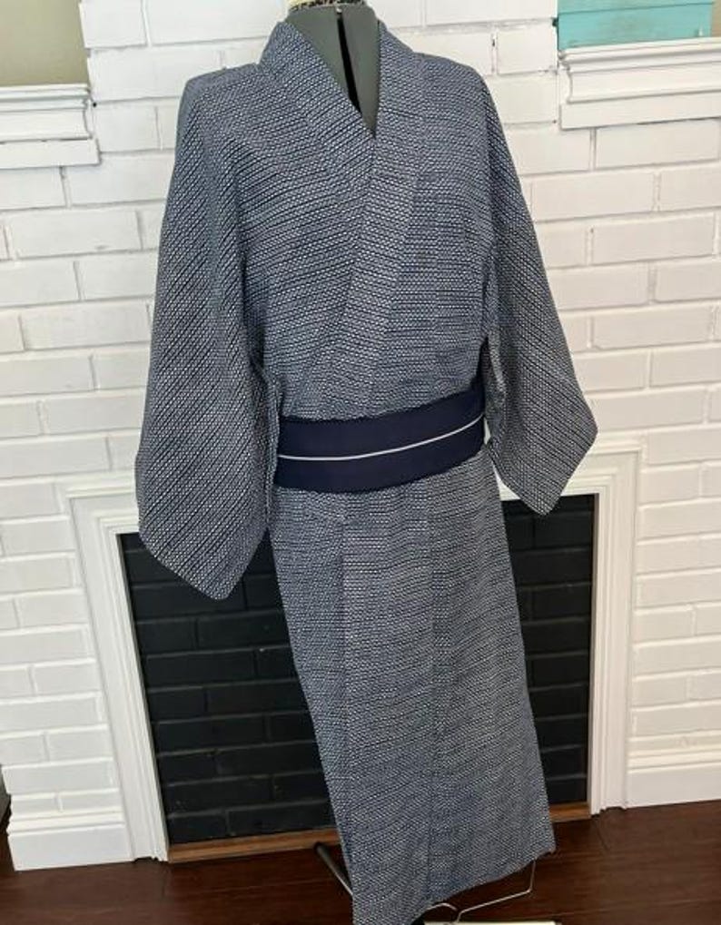 Może przedstawiać: Kimono w ciemnoniebiesko-biały wz&oacute;r z drobną geometryczną teksturą na całej tkaninie. Ubi&oacute;r posiada szerokie rękawy, dekolt w serek oraz jednolity ciemnogranatowy pas obi zawiązany w talii z cienkim białym poziomym paskiem.