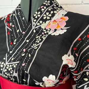 May include: A black kimono featuring a pattern of white and pink cherry blossoms, red petals, and diagonal grey stripes. The garment has a wrap-style front with a wide collar and is cinched at the waist with a solid red obi sash.