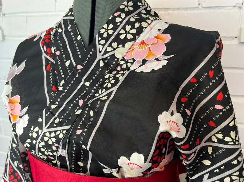 May include: A black kimono featuring a pattern of white and pink cherry blossoms, red petals, and diagonal grey stripes. The garment has a wrap-style front with a wide collar and is cinched at the waist with a solid red obi sash.