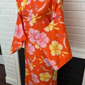 Może przedstawiać: Jaskrawopomarańczowe kimono z powtarzającym się kwiatowym wzorem w jasnor&oacute;żowe i ż&oacute;łte kwiaty. Ubi&oacute;r posiada szerokie, luźne rękawy oraz zapięcie typu kopertowego z przodu, zaprezentowane na manekinie, aby ukazać pełną długość i układ tkaniny.