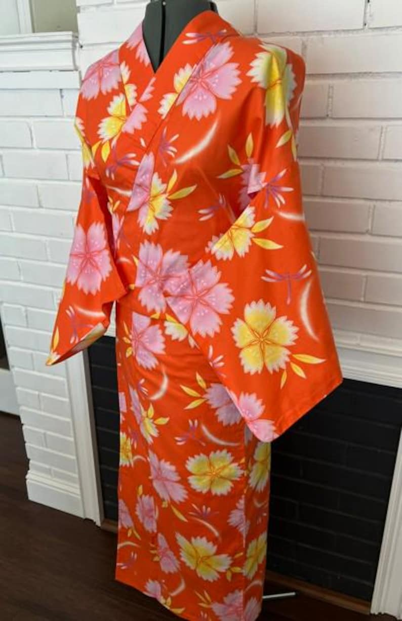 Może przedstawiać: Jaskrawopomarańczowe kimono z powtarzającym się kwiatowym wzorem w jasnor&oacute;żowe i ż&oacute;łte kwiaty. Ubi&oacute;r posiada szerokie, luźne rękawy oraz zapięcie typu kopertowego z przodu, zaprezentowane na manekinie, aby ukazać pełną długość i układ tkaniny.