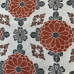 May include: A textile fabric featuring a repeating pattern of large, rust-orange chrysanthemum-like flowers with dark grey centres, interspersed with smaller grey four-petalled blossoms and geometric diamond shapes on a cream-coloured background.