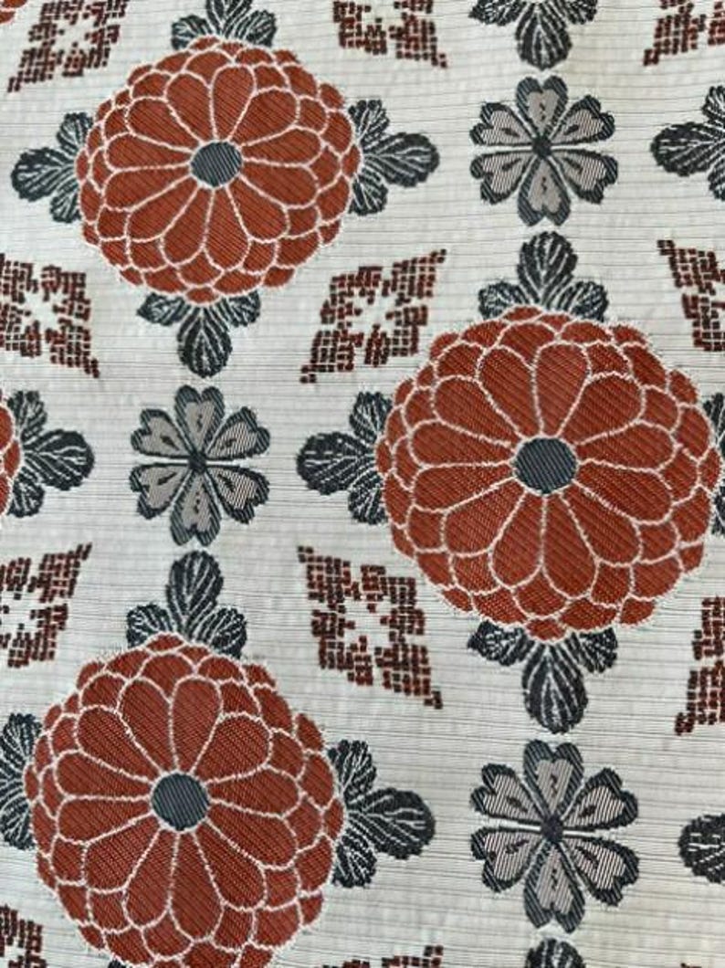 May include: A textile fabric featuring a repeating pattern of large, rust-orange chrysanthemum-like flowers with dark grey centres, interspersed with smaller grey four-petalled blossoms and geometric diamond shapes on a cream-coloured background.