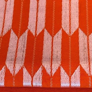 May include: A vibrant orange fabric featuring a repeating pattern of light pink, arrow-like shapes arranged in vertical columns. The textile displays a textured weave with thin, vertical lines running between the pale pink arrow motifs.