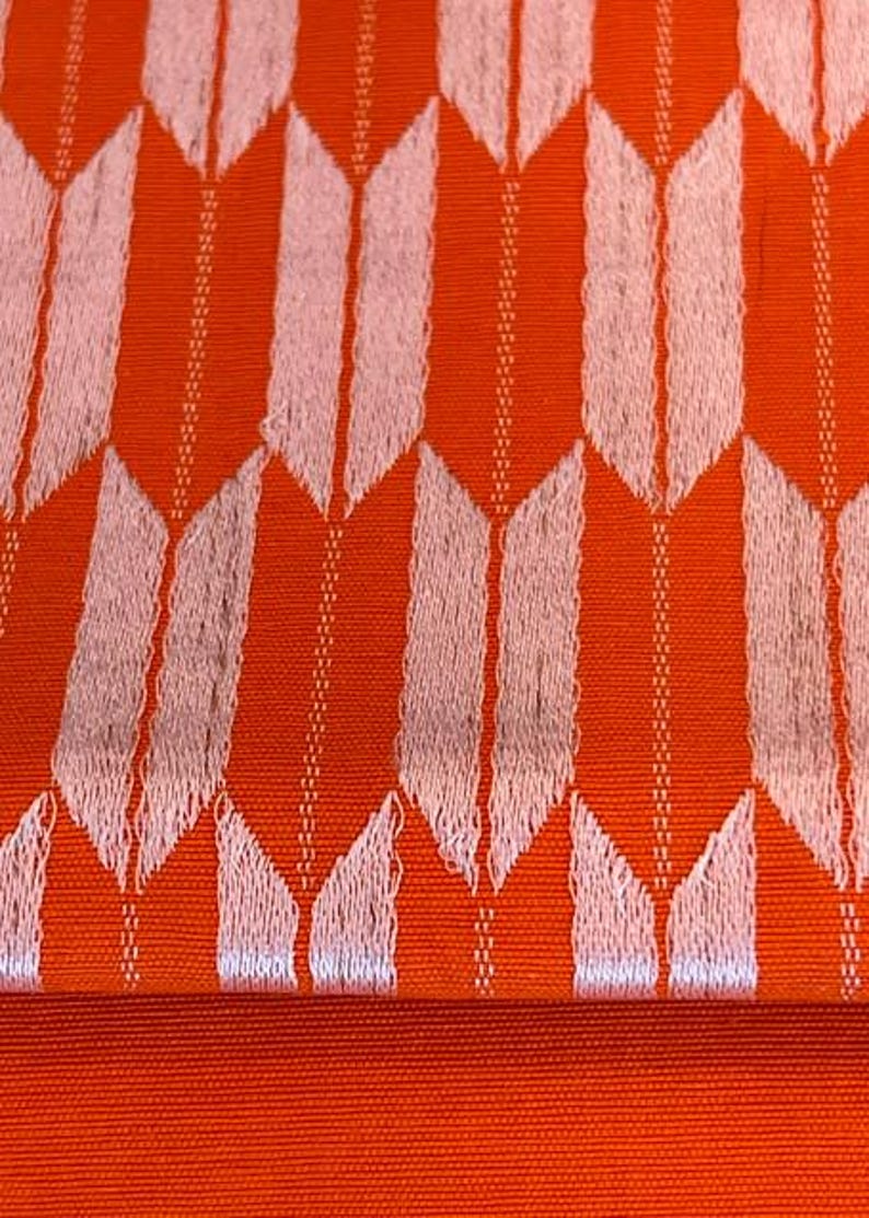 May include: A vibrant orange fabric featuring a repeating pattern of light pink, arrow-like shapes arranged in vertical columns. The textile displays a textured weave with thin, vertical lines running between the pale pink arrow motifs.
